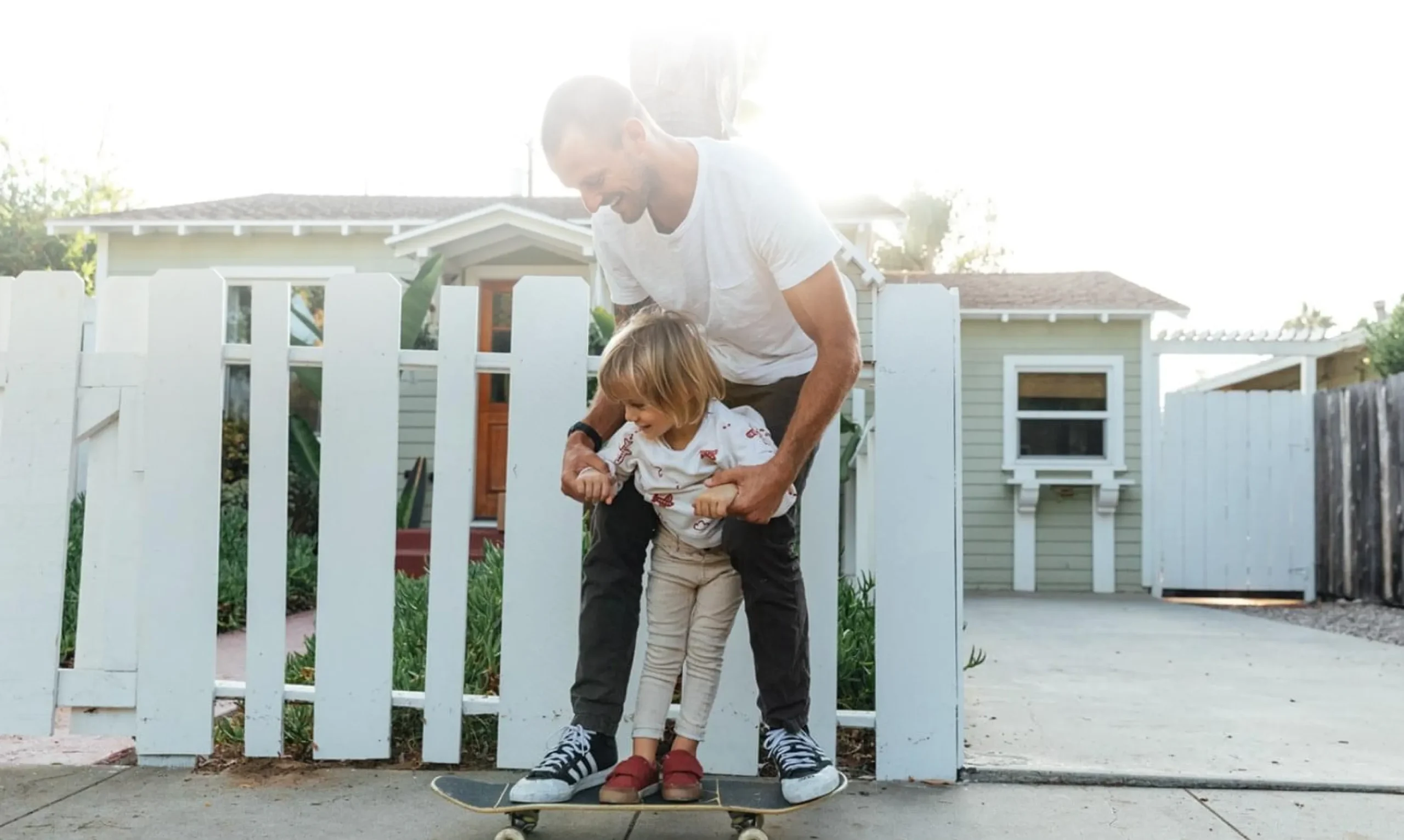 Father teaching child to skateboard by white picket fence.